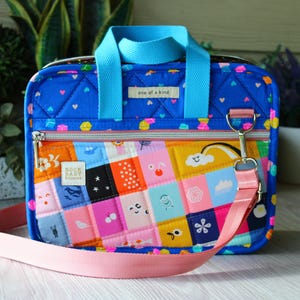 May include: A blue quilted bag with a light blue handle and a pink shoulder strap. The bag features a colorful patchwork design with various shapes and illustrations. The text "one of a kind" is on the top of the bag. The bag has a zipper closure.