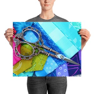 May include: A pair of silver sewing scissors with ornate floral handles rests on a colorful patchwork of fabric. The fabric features various shades of blue, green, pink, and purple, creating a vibrant and textured background.