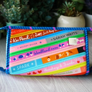 May include: A colorful, rectangular fabric bag with a blue handle. The bag features a patchwork design with various text elements, including "Koi Pond," "Woodland Park," and "Spark." The bag is trimmed with blue piping and has a small tag that says "Sarah Watts."