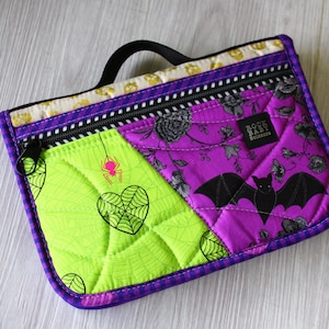 Könnte beinhalten: Gesteppte Halloween-Tasche mit schwarzem Griff. Die Tasche hat eine limettengrüne Platte mit Spinnennetz- und Herzdesign und eine lila Platte mit Fledermaus- und Blumenmuster. Ein schwarzer Reißverschluss und lila Zierleisten vervollständigen das Design.