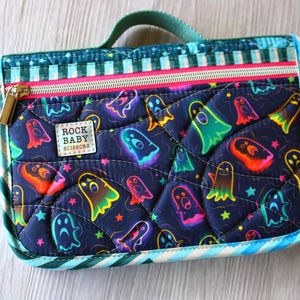 May include: A quilted rectangular zippered pouch featuring a dark blue fabric with a colorful ghost and star pattern. The pouch has a teal handle, a bright pink zipper, and a white rectangular label with the text ROCK BABY SCISSORS.