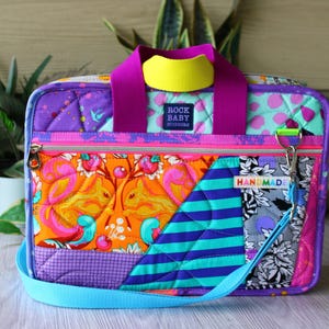 May include: A rectangular quilted bag featuring a vibrant patchwork design with orange squirrel motifs, teal stripes, and floral patterns. It has purple handles, a yellow accent, a front zipper, and a tag reading ROCK BABY SCISSORS and HANDMADE.