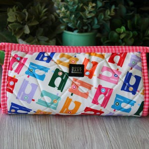 May include: A quilted cylindrical pouch featuring a white background with a repeating pattern of colorful sewing machines in red, orange, green, blue, and purple. It has a pink and white gingham trim, two side handles, and a black label reading Rock Baby Scissors.