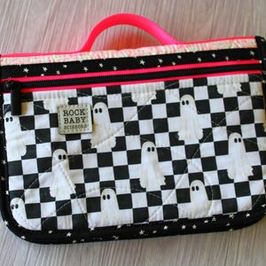 May include: A rectangular quilted zipper pouch featuring a black and white checkered pattern with white ghost illustrations. The pouch has a neon pink handle, a black zipper, and a small grey label with the text ROCK BABY SCISSORS.