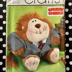 May include: Simplicity crafts pattern featuring a plush lion wearing a black and white houndstooth blazer and a red tie. The lion has a brown mane, a pink face, and the words "CHUBBY CHEEKS" are printed on the pattern.
