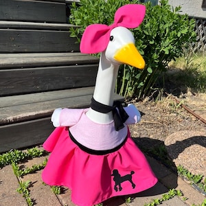 May include: A decorative goose figurine wearing a pink poodle skirt, a pink top, and a black bow tie. The goose has a white head, a yellow beak, and a large pink bow. The skirt has a black poodle silhouette.