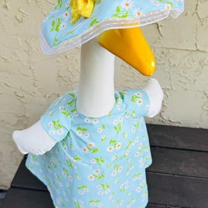 Pode incluir: Uma figura de ganso caprichosa vestindo um vestido azul claro e um chap&eacute;u combinando, ambos adornados com uma estampa de margaridas. O chap&eacute;u apresenta um la&ccedil;o amarelo com bolinhas brancas e uma borda de renda branca. O ganso tem um bico amarelo.