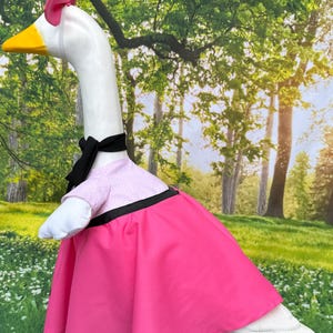 May include: A white goose costume with a pink dress, black bow, and pink bow on its head. The goose has a yellow beak and is set against a backdrop of trees and grass. The costume includes a pink dress with a black trim.