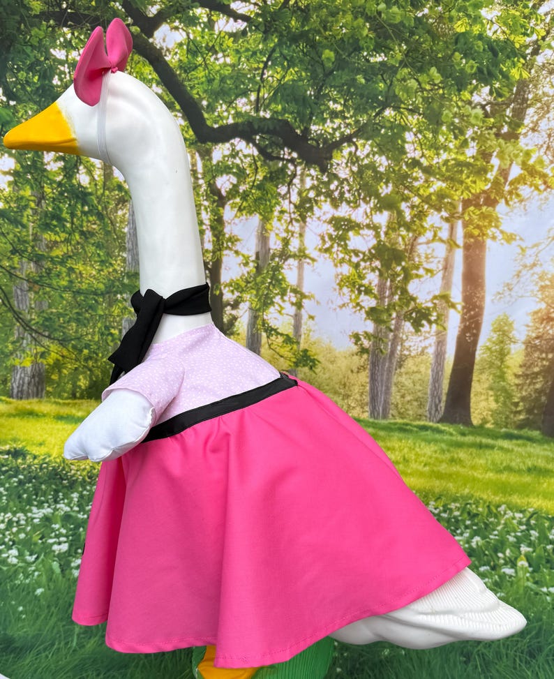 May include: A white goose costume with a pink dress, black bow, and pink bow on its head. The goose has a yellow beak and is set against a backdrop of trees and grass. The costume includes a pink dress with a black trim.