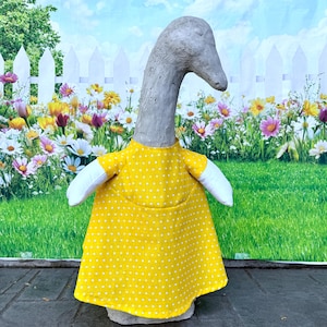 Könnte beinhalten: Eine Beton-Gans-Statue, die ein gelbes Kleid mit weißen Punkten und weißen Ärmeln trägt. Die Gans hat einen langen Hals und einen grauen Kopf. Der Hintergrund zeigt einen weißen Zaun und einen bunten Blumengarten.