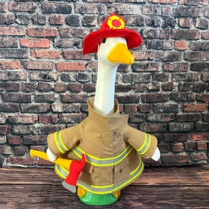 May include: A plush goose dressed as a firefighter, wearing a red helmet with a yellow emblem, a tan jacket with reflective stripes, and holding a yellow axe with a red handle. The goose has a white neck and a yellow beak.