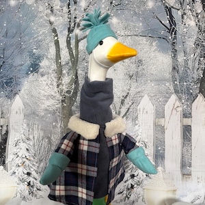 Puede incluir: Una caprichosa figura de ganso vestida para el invierno. El ganso lleva un gorro turquesa, una bufanda gris, una chaqueta a cuadros con cuello de piel sintética y mitones turquesas. El fondo presenta una escena invernal nevada con una valla blanca.