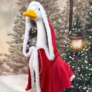 May include: A white goose statue adorned with a red and white Christmas cape and hood. The hood is trimmed with white faux fur and lined with a festive print. The goose has a yellow beak and is set against a backdrop of Christmas trees.