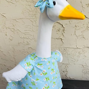 Pode incluir: Uma estatueta de ganso branco com bico amarelo e um vestido azul claro adornado com margaridas brancas e folhas verdes. O ganso usa uma faixa de cabe&ccedil;a de flor combinando. A estatueta est&aacute; em uma superf&iacute;cie escura.