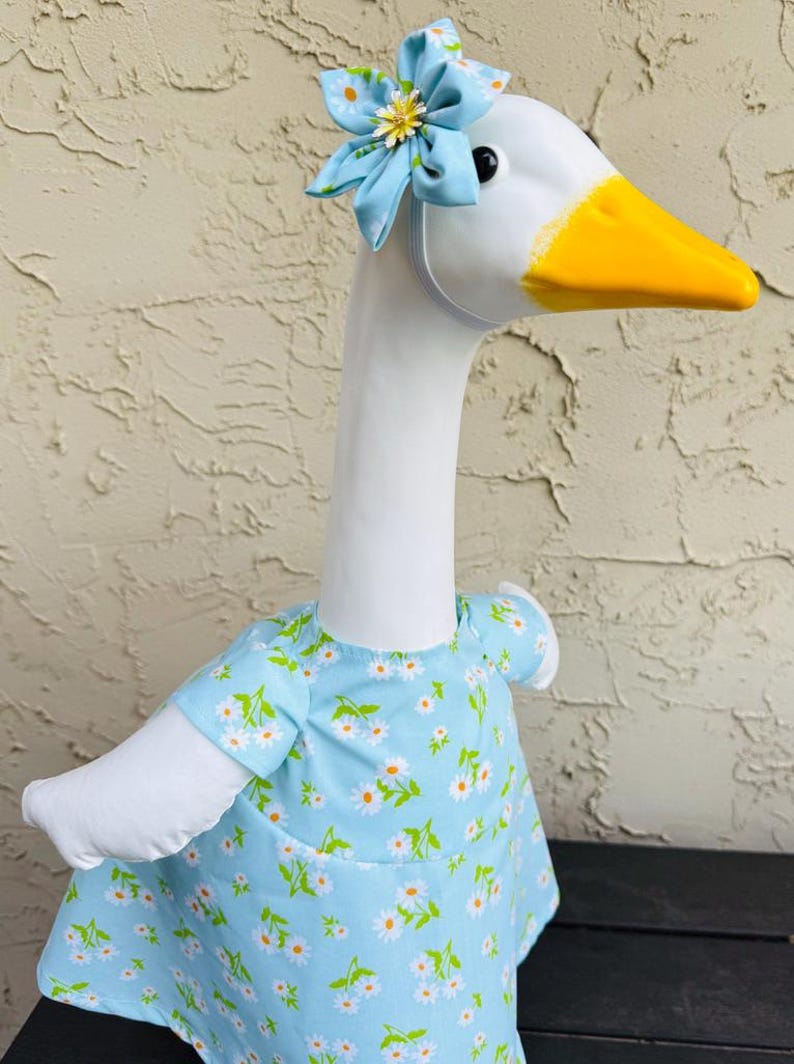 Pode incluir: Uma estatueta de ganso branco com bico amarelo e um vestido azul claro adornado com margaridas brancas e folhas verdes. O ganso usa uma faixa de cabe&ccedil;a de flor combinando. A estatueta est&aacute; em uma superf&iacute;cie escura.
