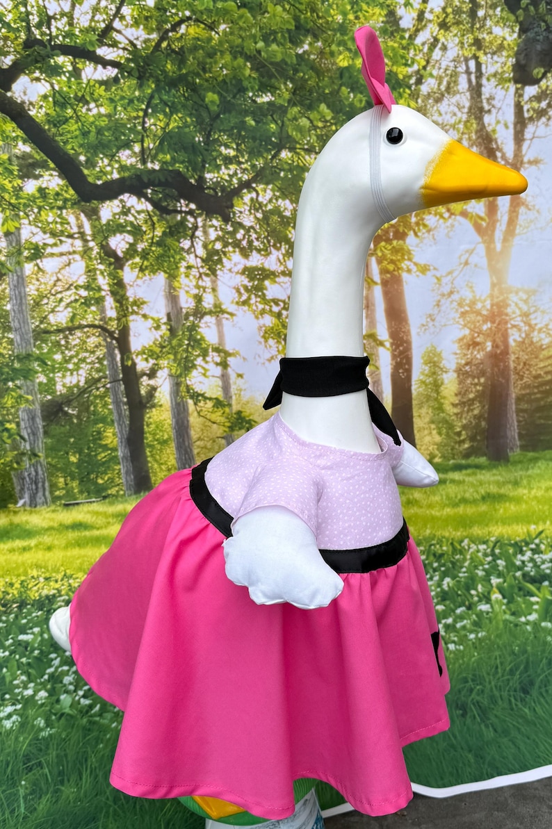 May include: A goose costume with a white head, yellow beak, and pink feather. The goose wears a pink dress with black trim and a black necktie. The background is a forest scene.