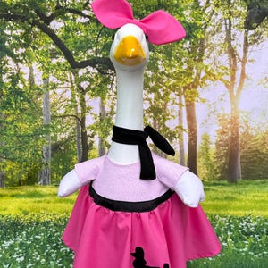 May include: A goose figurine dressed in a pink poodle skirt, a pink top with white polka dots, and a black scarf. The goose wears a large pink bow on its head. The skirt has a black poodle silhouette.