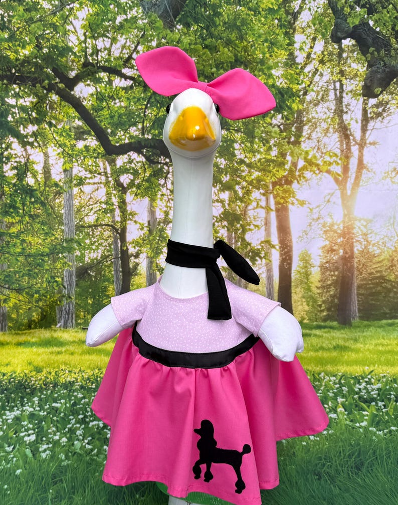 May include: A goose figurine dressed in a pink poodle skirt, a pink top with white polka dots, and a black scarf. The goose wears a large pink bow on its head. The skirt has a black poodle silhouette.