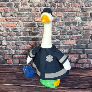 May include: A plush goose wearing a navy blue firefighter uniform with reflective stripes and a black cap. The goose has a yellow beak and is holding a blue bag. The background is a brick wall.
