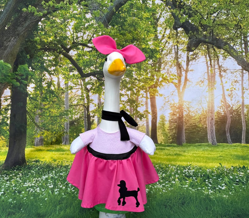 May include: A goose figurine dressed in a pink outfit with a black poodle silhouette. The goose wears a pink bow, a black scarf, and a pink dress with a black trim. The background is a sunny outdoor scene with trees and grass.