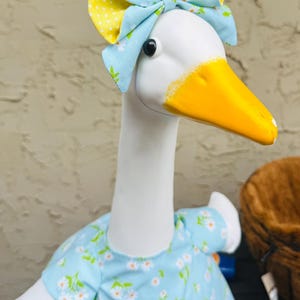 Pode incluir: Uma estatueta de ganso branco com bico amarelo e olhos pretos. Usa um vestido azul claro com um padr&atilde;o de margaridas brancas e um la&ccedil;o a condizer com bolinhas amarelas. O ganso &eacute; um item decorativo.