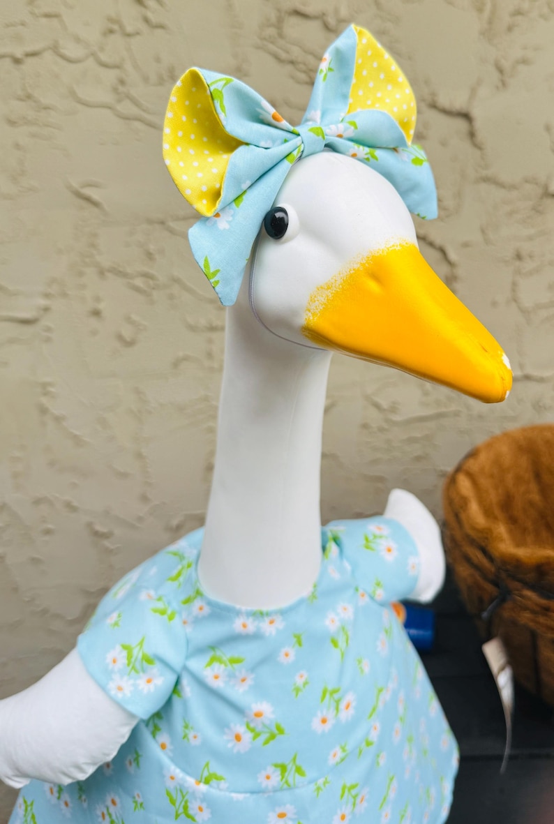 Pode incluir: Uma estatueta de ganso branco com bico amarelo e olhos pretos. Usa um vestido azul claro com um padr&atilde;o de margaridas brancas e um la&ccedil;o a condizer com bolinhas amarelas. O ganso &eacute; um item decorativo.