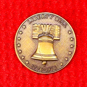 May include: A circular bronze-toned commemorative coin featuring a raised relief of the Liberty Bell in the center. The text LIBERTY BELL is arched across the top, and the dates 1776-1976 are at the bottom, surrounded by a ring of thirteen stars.