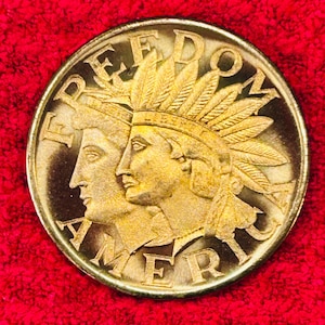 May include: A gold-colored coin with the words "FREEDOM AMERICA" encircling two profile portraits. One portrait wears a feathered headdress. The coin is set against a red background.