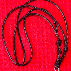 May include: A black braided cord necklace featuring a series of adjustable sliding knots and a small silver-toned metal ring at the bottom end, designed for holding pendants or other small decorative items securely.