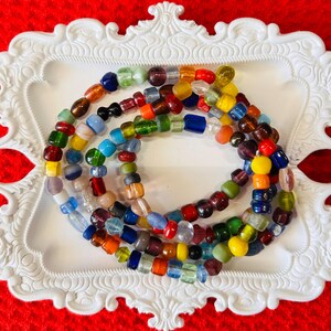 May include: A multi-strand necklace composed of numerous small, irregularly shaped glass beads in a wide variety of colors including red, blue, green, yellow, orange, purple, and clear, arranged in a circular pattern on a white decorative tray.