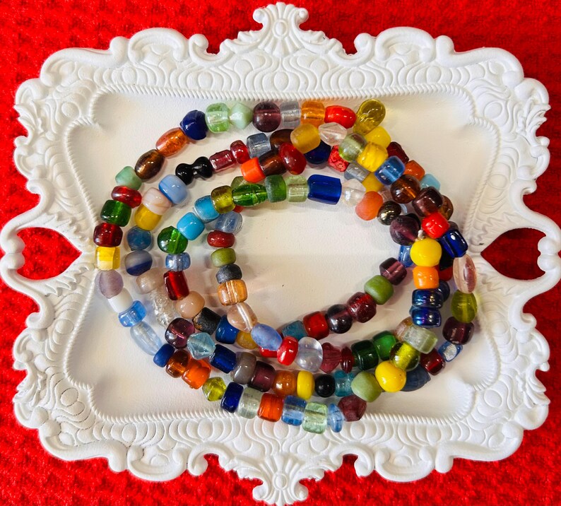May include: A multi-strand necklace composed of numerous small, irregularly shaped glass beads in a wide variety of colors including red, blue, green, yellow, orange, purple, and clear, arranged in a circular pattern on a white decorative tray.