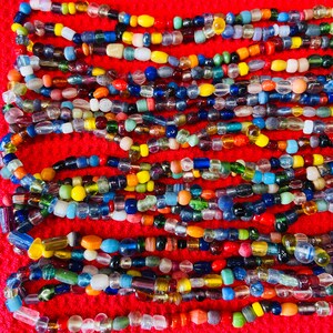 May include: A collection of multi-colored glass beads arranged in horizontal rows. The beads vary in shape, size, and opacity, featuring a spectrum of colors including red, blue, yellow, green, orange, white, and clear glass, all set against a red background.