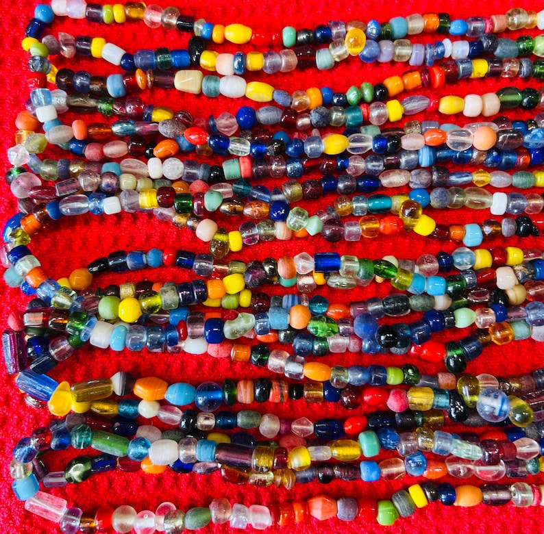 May include: A collection of multi-colored glass beads arranged in horizontal rows. The beads vary in shape, size, and opacity, featuring a spectrum of colors including red, blue, yellow, green, orange, white, and clear glass, all set against a red background.