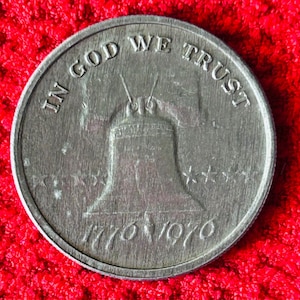 May include: A silver coin depicting the Liberty Bell, with the inscriptions "IN GOD WE TRUST" and "1776 1976". The coin is set against a red background.