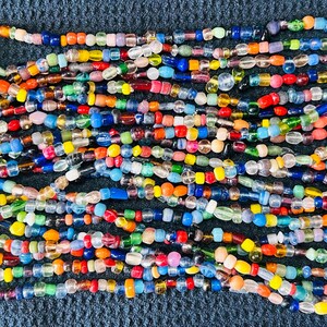 May include: A long, multi-strand necklace composed of numerous small, irregular glass beads in a vibrant array of colors including red, blue, yellow, green, orange, and clear. The beads are strung together in a dense, layered, and chaotic pattern.