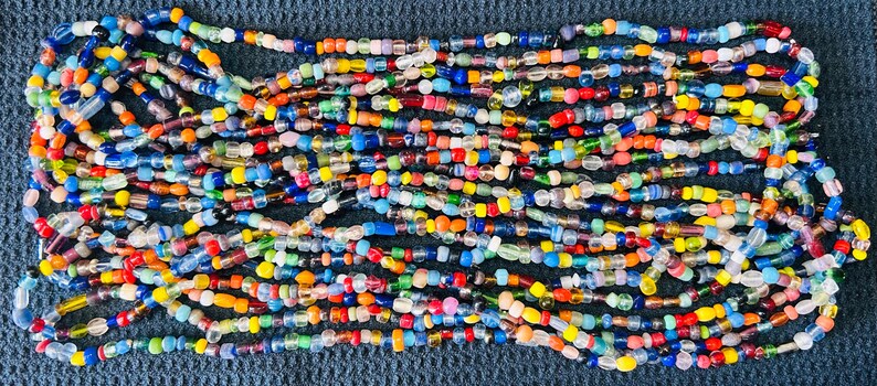 May include: A long, multi-strand necklace composed of numerous small, irregular glass beads in a vibrant array of colors including red, blue, yellow, green, orange, and clear. The beads are strung together in a dense, layered, and chaotic pattern.