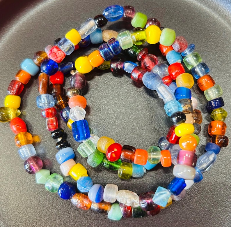 May include: A circular strand of multi-colored glass beads featuring a variety of shapes and sizes. The beads display a vibrant spectrum of colors including red, blue, yellow, green, orange, and clear tones, arranged in a continuous loop pattern.