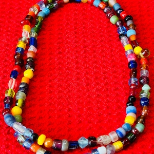 May include: A long necklace composed of a single strand of small, multi-colored glass beads. The beads feature a variety of translucent, opaque, and clear finishes in shades of blue, yellow, red, green, orange, white, black, and purple.