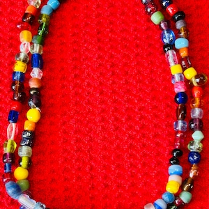 May include: A long necklace composed of a single strand of multi-colored glass beads in various shapes and sizes. The beads feature a wide spectrum of colors including red, blue, yellow, green, orange, purple, black, white, and clear glass.
