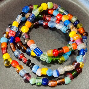 May include: A multi-strand necklace composed of numerous small, translucent and opaque glass beads in a vibrant array of colors including red, blue, yellow, green, orange, and clear. The beads vary in shape, featuring round, cylindrical, and faceted forms.