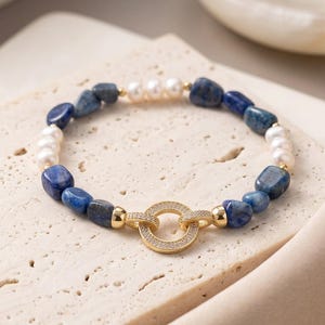 Puede incluir: Una pulsera de cuentas con segmentos alternos de perlas blancas lisas y piedras de lapis lázuli azules pulidas. La pulsera se asegura con un cierre circular dorado incrustado con pequeñas piedras claras, conectado por dos cuentas doradas pulidas.