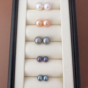 Puede incluir: Una exhibición de seis pares de pendientes de perlas en una caja de joyería negra. Las perlas son de varios colores, incluyendo blanco, melocotón, gris, púrpura y verde azulado. Los pendientes están dispuestos en filas sobre un fondo crema.