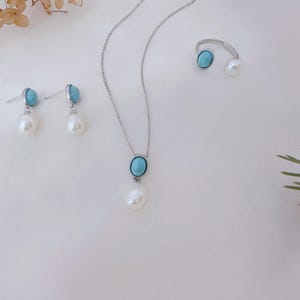 May include: A jewellery set featuring a silver necklace, earrings, and ring. Each piece has a light blue oval stone and a white teardrop-shaped pearl. The jewellery is displayed on a white surface.