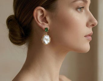 May include: Gold-toned earrings featuring a rectangular green gemstone at the top and a large, irregular white pearl pendant. The pearl is encased in a gold wire frame. The earrings are worn on a person's ear.