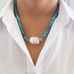 May include: A beaded necklace featuring round, mottled turquoise-colored stones and a single, large, irregular white baroque pearl centered at the front. The necklace is worn against a crisp, white collared shirt with a visible button placket.