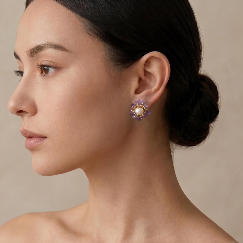 May include: A close-up of a woman wearing a pair of flower-shaped earrings. Each earring features a central pearl surrounded by small purple gemstones and gold-colored metal accents. The earrings are set against the woman's ear and skin.