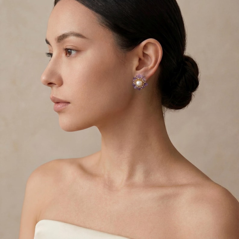 May include: A close-up of a woman wearing a pair of floral-shaped earrings. Each earring features a central pearl surrounded by small purple gemstones and gold-colored metal detailing. The earrings are set against the woman's ear.