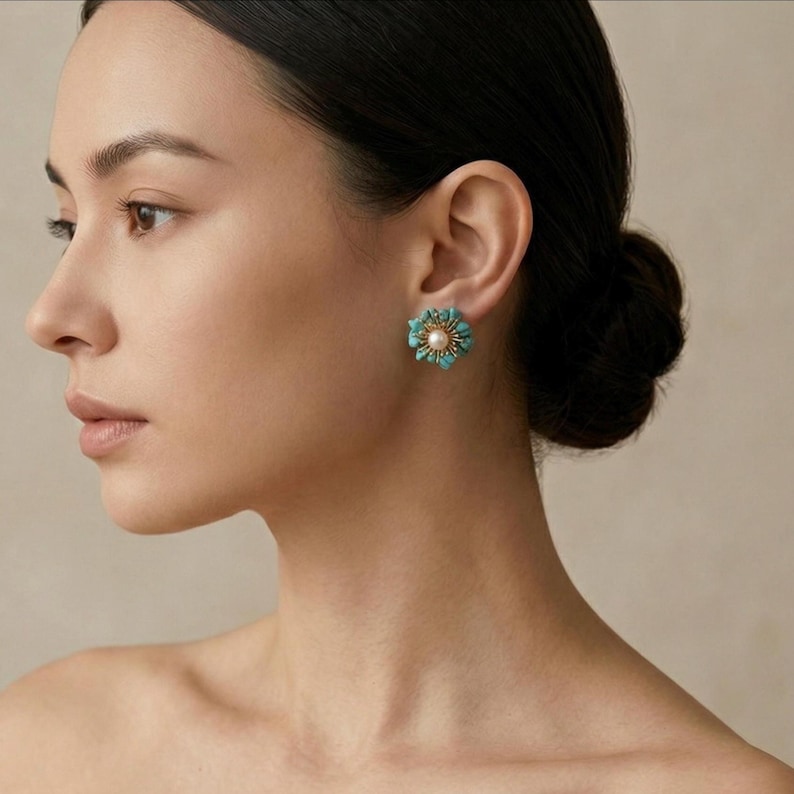 May include: A close-up of a turquoise and gold flower-shaped earring with a pearl center. The earring is worn on the ear of a person with dark hair pulled back. The earring has a detailed design.