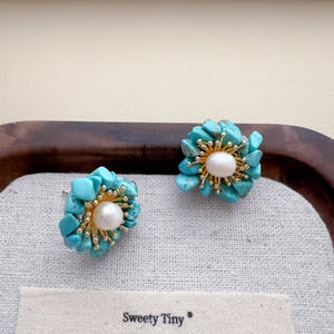 May include: A pair of flower-shaped stud earrings. Each earring features a central white pearl surrounded by turquoise-colored stones and gold-colored metal accents. The earrings are displayed on a white fabric with the text "Sweety Tiny".