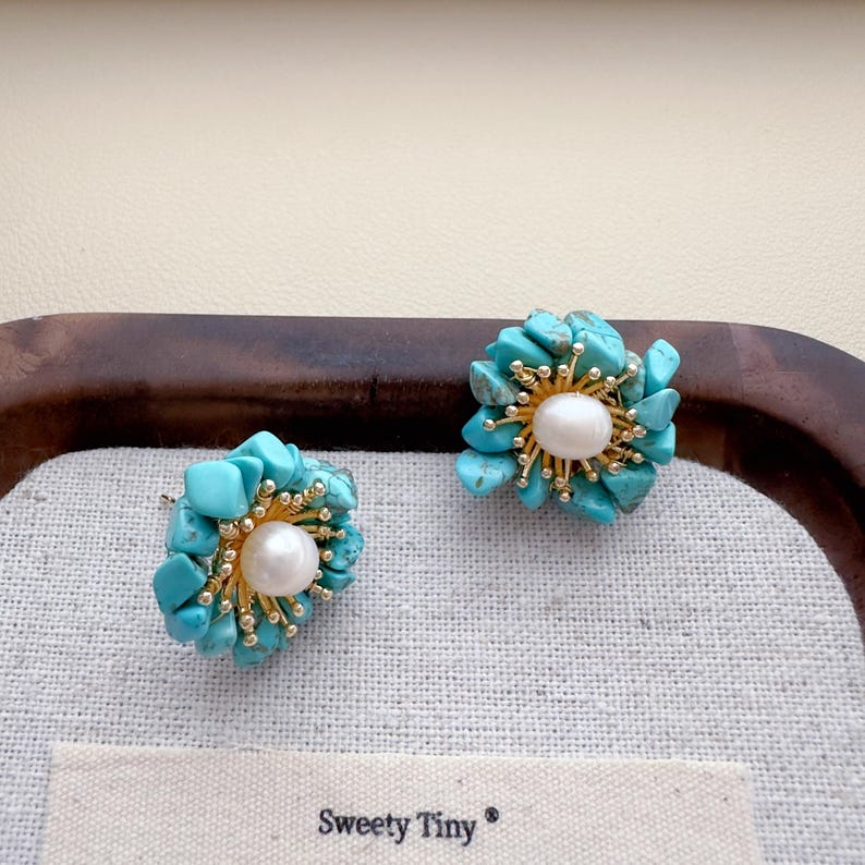 May include: A pair of flower-shaped stud earrings. Each earring features a central white pearl surrounded by turquoise-colored stones and gold-colored metal accents. The earrings are displayed on a white fabric with the text "Sweety Tiny".